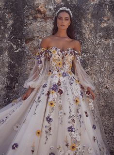 a woman in a white dress with flowers on it standing next to a stone wall