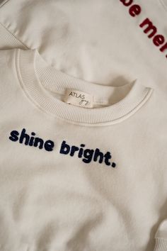 Two cream-colored toddler outfits are laid flat on a light surface. The left outfit has "be merry." written in red on the sweatshirt, while the right outfit reads "shine bright." in navy blue. Both sets include matching ribbed pants. Special Moments, Graphic Crewneck, Shine Bright, Coconut Milk, Sale Items