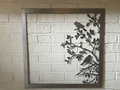 a metal bird on a branch with leaves and flowers in a square frame against a white brick wall