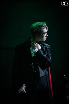 a man with green hair standing in the dark wearing a black suit and red cape