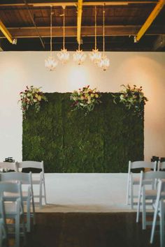 chairs are set up in front of a moss wall with flowers on it and chandeliers hanging from the ceiling