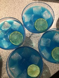 three glasses filled with blue liquid and limes on top of a granite countertop