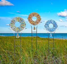 three metal sculptures are in the grass by the beach with blue skies and water behind them