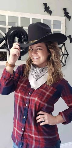 a woman wearing a red plaid shirt and black cowboy hat with her hands on her hip