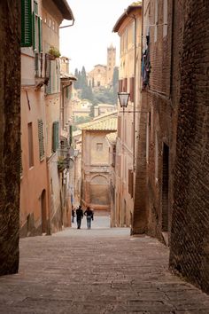 two people walking down an alley way in the middle of town with buildings on either side