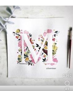 the letter m is painted with watercolors and surrounded by flowers on a piece of paper