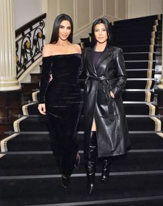 two women standing next to each other in front of stairs wearing black dresses and coats