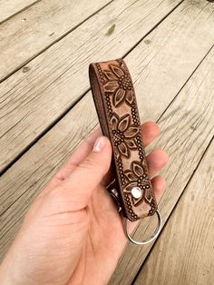 a hand holding a brown leather keychain on top of a wooden deck with flowers