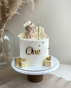 a white cake with gold decorations and a small teddy bear on top that says one