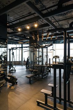 an empty gym with lots of machines and lights on the ceiling in front of large windows