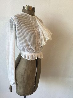 "Vintage Victorian white cotton net and lace blouse with long sleeves and a very large flouncy ruffle coming off a crochet placard down the front. The net has pintuck creating a ribbon effect The sleeves each have two functioning buttons and a small trim of lace. The waist is gathered to create a ruffle around and is backed with ribbon. The whole blouse is lined with net. The blouse buttons down the back with a covered placard to hide the buttons. Single hook and eye at waist. Simple crew neck. White Lace Top With Ruffles, Lace Top With Ruffles For Daywear, Fitted Victorian Ruffle Top, White Victorian Style Top With Lace Work, White Long Sleeve Lace Top With Lace Cuffs, Victorian Ruffle Blouse For Daywear, Fitted Cotton Lace Top With Ruffles, Victorian White Long Sleeve Blouse, White Victorian Long Sleeve Blouse