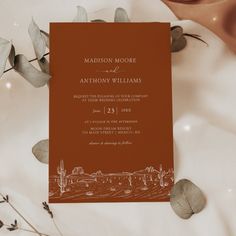 an orange and white wedding card on top of a table next to some flowers with leaves