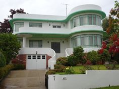 a large white house with green trim on the front and side windows, surrounded by greenery