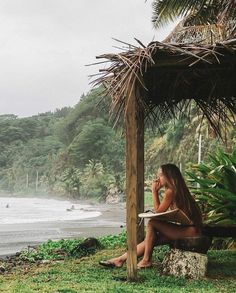 a woman sitting on top of a wooden bench next to the ocean under a thatched roof