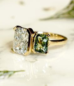 two green and white diamond engagement rings sitting on top of a marble surface with greenery in the background