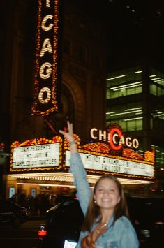 a woman standing in front of a theater holding her hand up to the sky and smiling