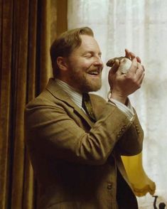 a man in a brown suit holding a small object up to his face while standing next to a window