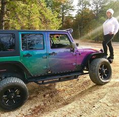 a man standing on top of a purple and green jeep parked in the dirt next to trees