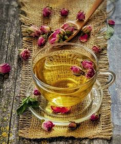 a glass cup filled with tea next to pink flowers on a wooden tablecloth and spoon