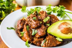 a white plate topped with meat and avocado next to cilantro on a wooden table