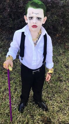 a young boy dressed as the joker with green hair and makeup holding a purple stick