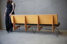 a woman standing next to a bench made out of plywood and metal legs, in front of a concrete wall