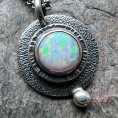 A simulated opal cabochon has been set in sterling and fine silver. I have made this unique necklace by sawing, soldering, texturing, and finishing the design entirely by hand. Pendant hangs from a sterling silver chain. The silver has been oxidized and polished to create a patina finish. Be sure to see all photos and video for size and color reference. Cabochon Size: About 8mm The necklace features a lobster claw clasp and a 3-inch extender allowing it to be slightly adjustable. Select your len Silver Opal Cabochon Necklace, Handmade Opal Round Pendant Necklace, Opal Cabochon Necklaces, Unique Oval Opal Necklaces, Handmade Opal Necklace, Opal Cabochon Round Pendant Necklace, Opal Cabochon Round Pendant Necklaces, Unique Ethiopian Opal Round Jewelry, Artisan Silver Opal Jewelry