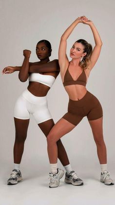 two women in brown and white bodysuits pose for the camera, one with her arms up