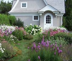 a house surrounded by flowers and greenery in front of the door is white with purple trim