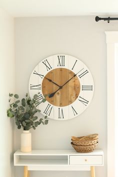 a white clock with roman numerals on the face next to a potted plant