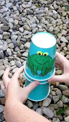 a person holding a blue cup with a green frog on it's side and rocks in the background