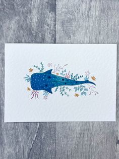 a card with a blue whale surrounded by leaves and flowers on a wooden surface,