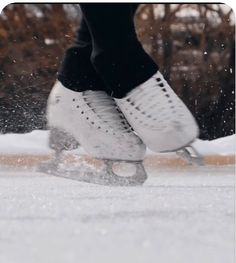 a person skating on an ice rink with snow falling off the ground and trees in the background