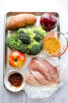 a tray filled with meat and vegetables on top of a white wooden table next to an apple