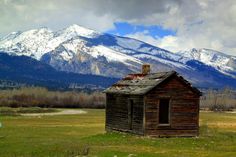 an old cabin in the middle of a field with mountains in the backgroud