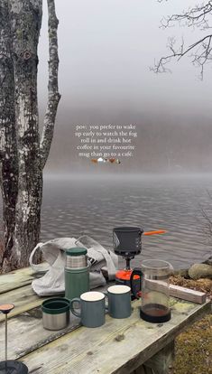 a picnic table with camping gear on it next to a lake and trees in the fog