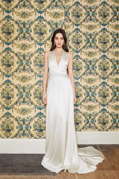 a woman standing in front of a wallpapered background wearing a long white dress