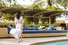 a woman in white dress walking by a swimming pool with blue couches and palm trees