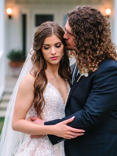 a bride and groom kissing in front of a white house on their wedding day with long curly hair