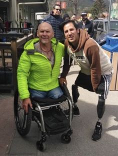 two men in wheelchairs standing next to each other on the street with people behind them