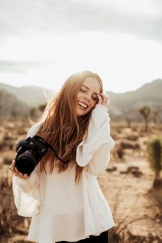 a woman holding a camera and smiling while standing in the middle of a desert area