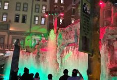 people are standing in front of a water fountain with lights on it at night time