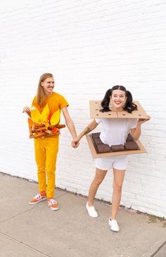 two women holding hands and standing in front of a white brick wall with an open cardboard box on their head