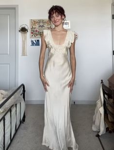 a woman in a white dress standing next to a bed and looking at the camera