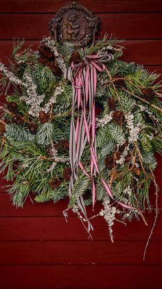 a christmas wreath with pine cones and pink ribbon hanging on a red wooden wall next to a plaque