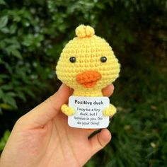 a hand holding a small yellow crocheted duck with a sign attached to it