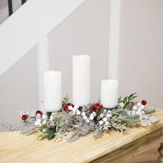 three white candles sitting on top of a wooden table with greenery and berries around it