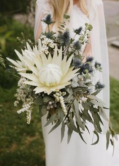 a woman in a white dress holding a bouquet of flowers and greenery on her wedding day