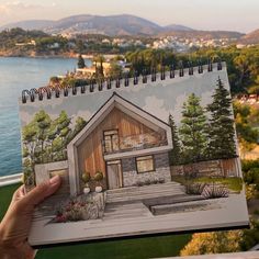 a person holding up a drawing of a house in front of some water and trees