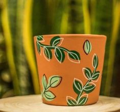 an orange cup with green leaves on it sitting on a wooden table next to a potted plant